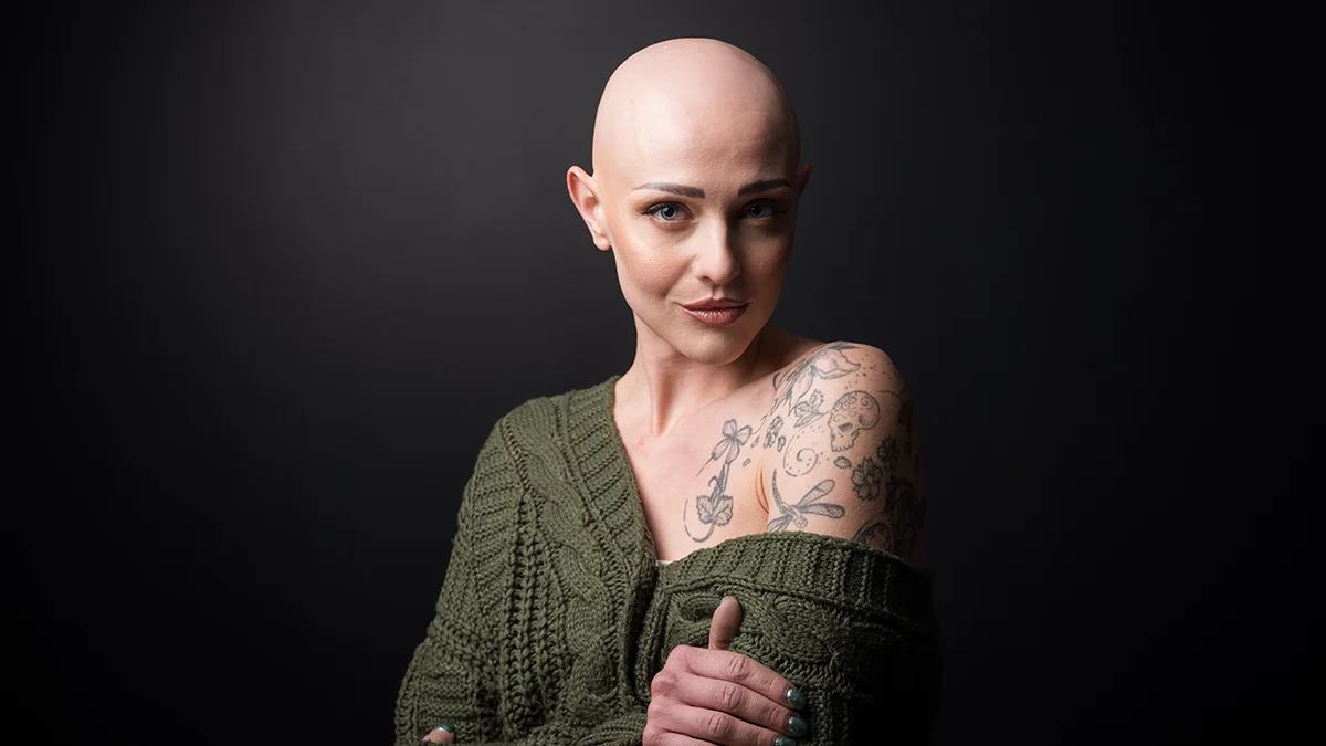 A beautiful woman with tatoos and alopecia, wearing a green off-shoulder sweater